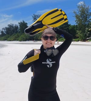 Elena smiles at the camera. She is standing on a bright white beach in a black wetsuit, and holding her yellow scuba fins above her head. 