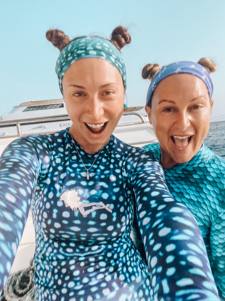 Two women divers smile at the camera wearing scuba headbands and with their hair in two small "space" buns on the top of their heads.