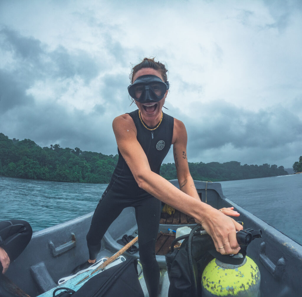 A woman stands in a small boat in the ocean with a lush green island behind her. She is smiling at the camera, wearing a scuba mask, and turning the valve on a scuba cylinder. 