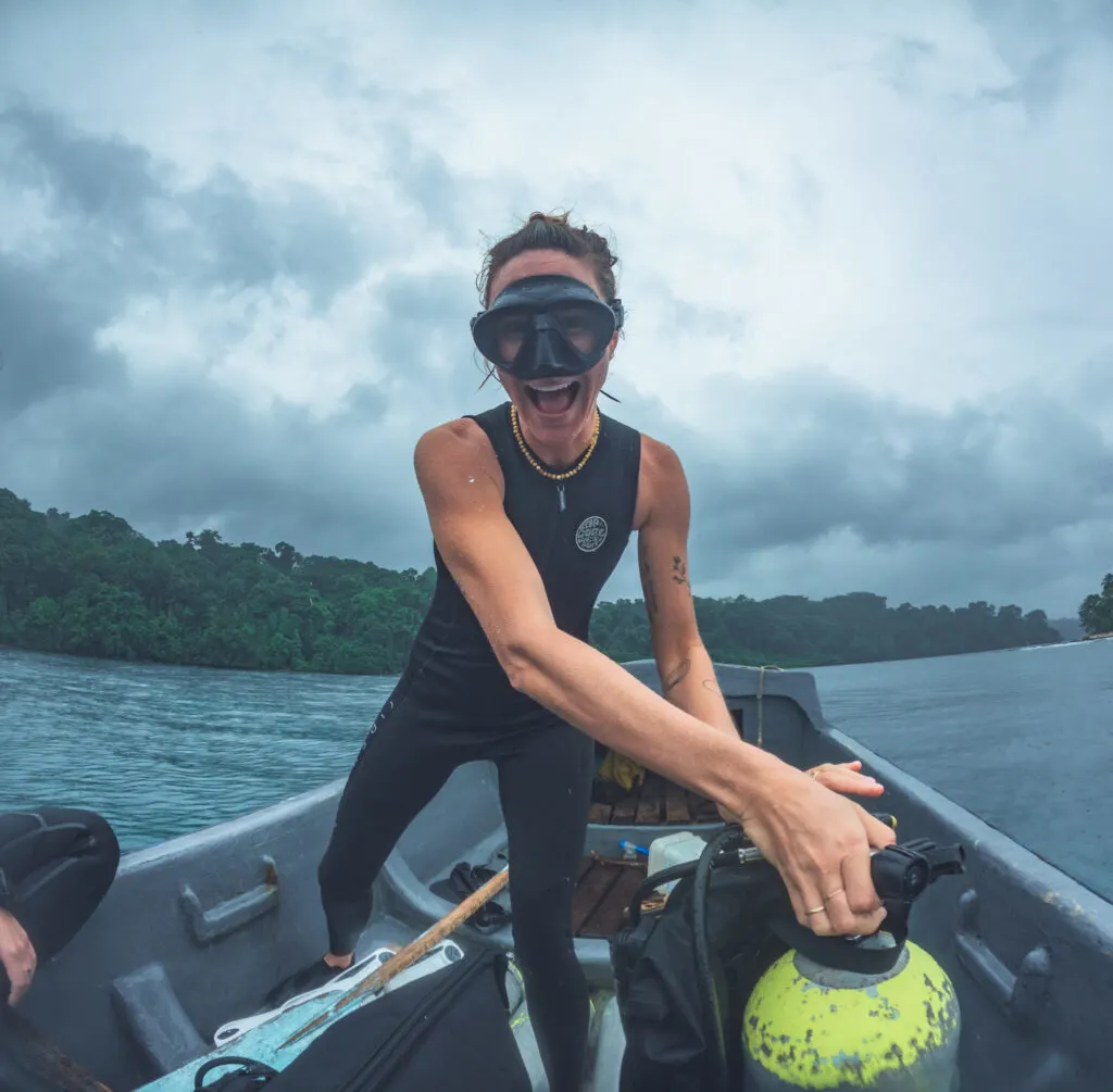 A woman stands in a small boat in the ocean with a lush green island behind her. She is smiling at the camera, wearing a scuba mask, and turning the valve on a scuba cylinder. 