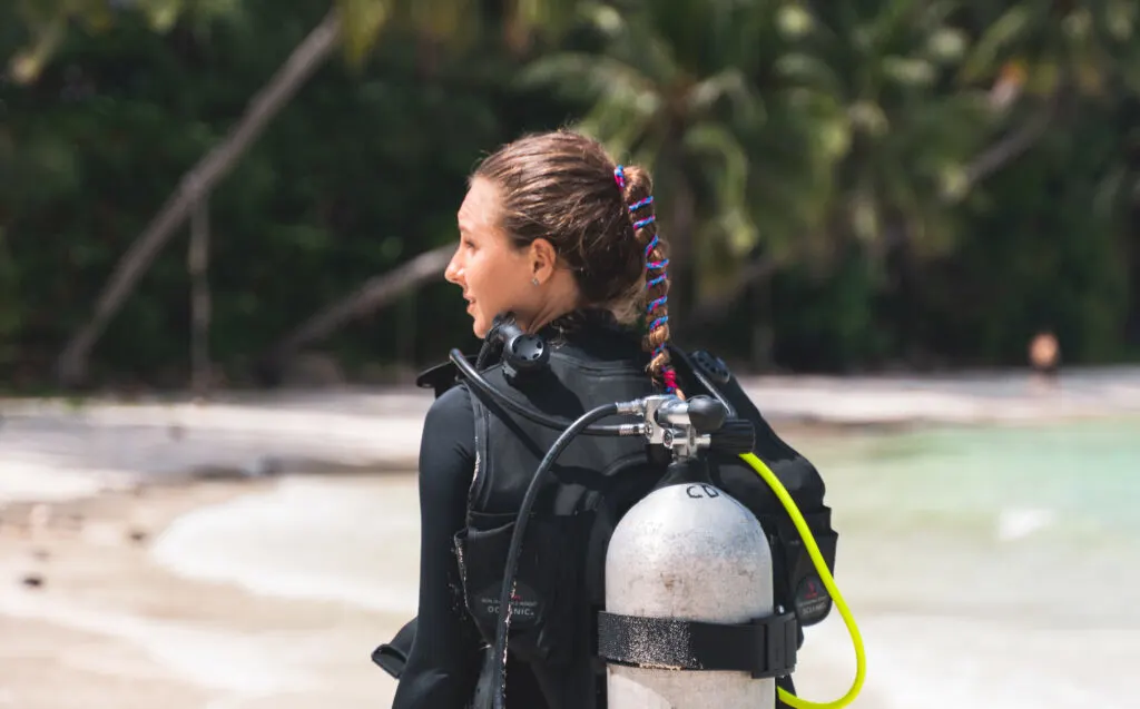 A scuba diver walks away from the camera on a white, sandy beach. Her long hair is tied up with a blue and pink hair tie.