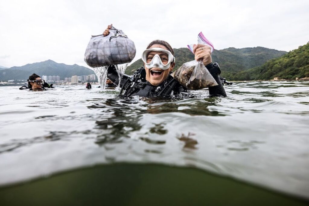 A woman scuba diver smiles at the surface of green-ish water. She is wearing a white scuba mask and holding two bags of shellfish, retrieved on a scientific dive. 