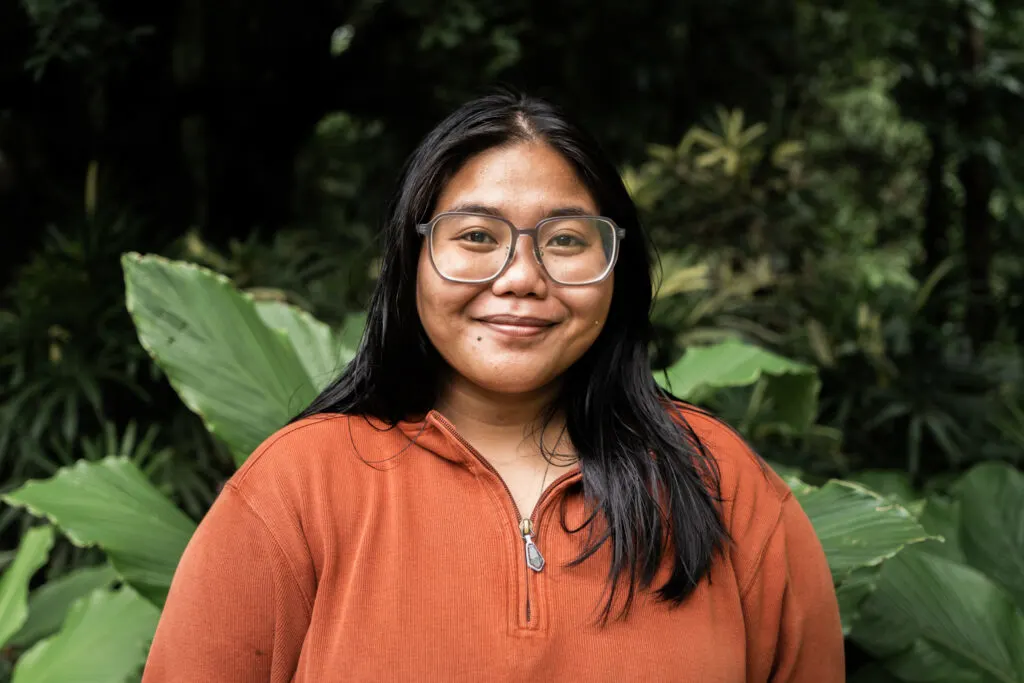 Bea Nodado smiles at the camera. She is standing in front of a leafy green backround, and she is wearing an orange zip-up top and large framed beige glasses.