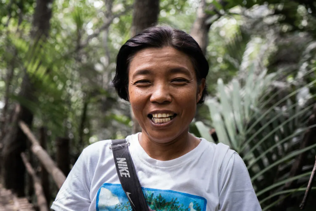 Evelyn smiles at the camera. She is stood in a jungle environment and wearing a white t-shirt with a blue image on it.