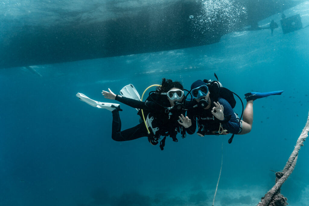 Two women scuba dive in deep blue water close to a boat. One gives an "okay" signal to the camera, the other holds her arms out.