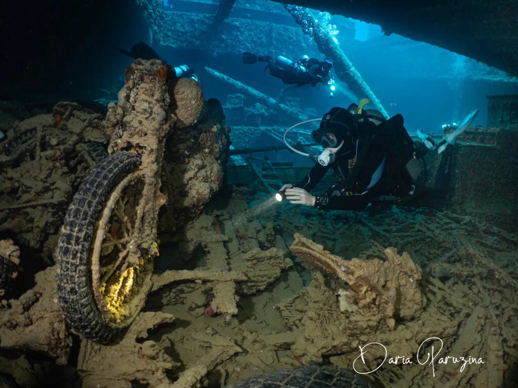 A woman scuba diver inside the Thistlegorm wreck in Egypt's Red Sea. She is pointing her dive light at a motorbike, and other divers swim in the open passage in the background. 