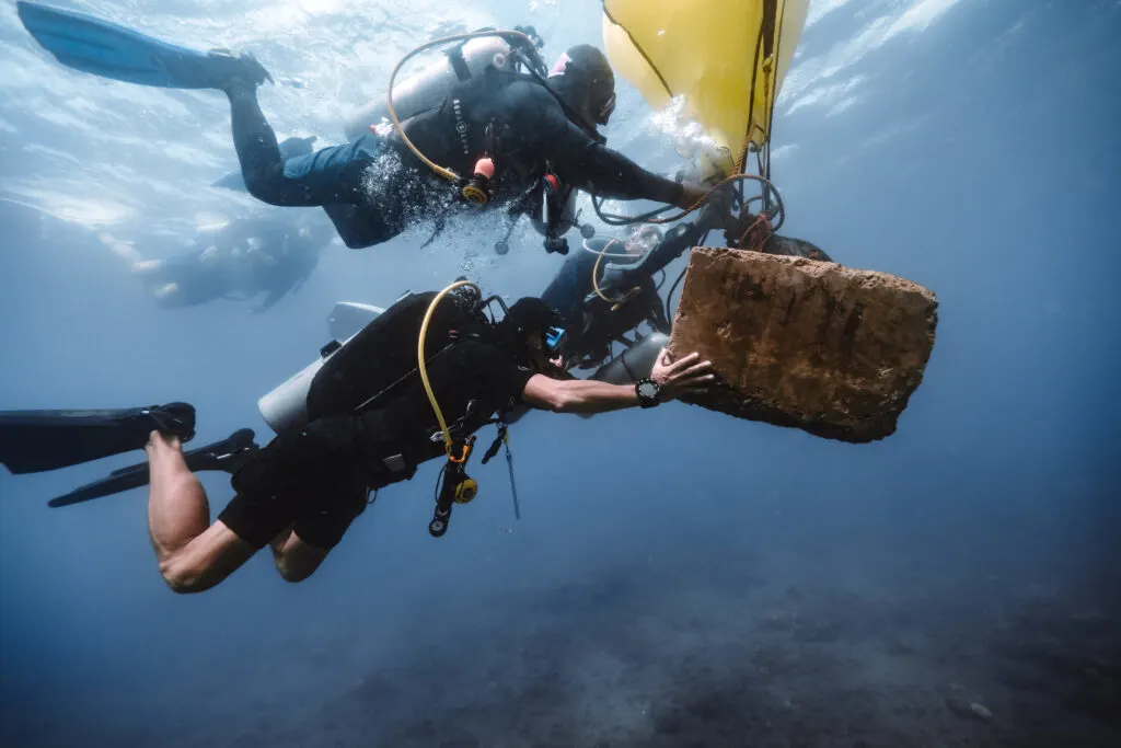 A group of scuba divers collaborate to send a yellow lift bag to the surface, raising a brown item with it.