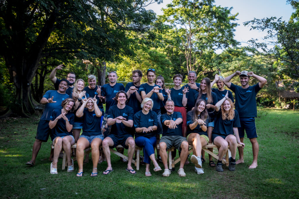 A large group of people smile and perform hand signals for the camera. They are wearing matching blue t-shirts and are in a lush, green clearing. 