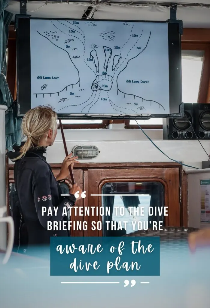 A blonde dive instructor wearing a black wetsuit points to a dive site map on a screen, she is giving a dive briefing.