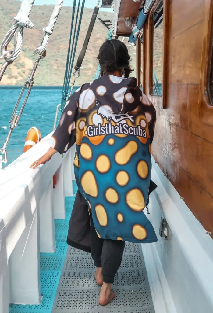 A woman scuba diver walks away from the camera on a large dive boat. She is wearing a microfibre poncho with a blue, white, and yellow print imitating a boxfish, with the Girls that Scuba logo of a woman diver silhouette.