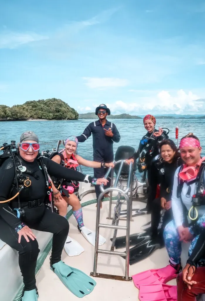 A group of women divers smile at the camera, with the boat captain smiling in the background. They are surrounded by bright blue water and idyllic small green islands. 