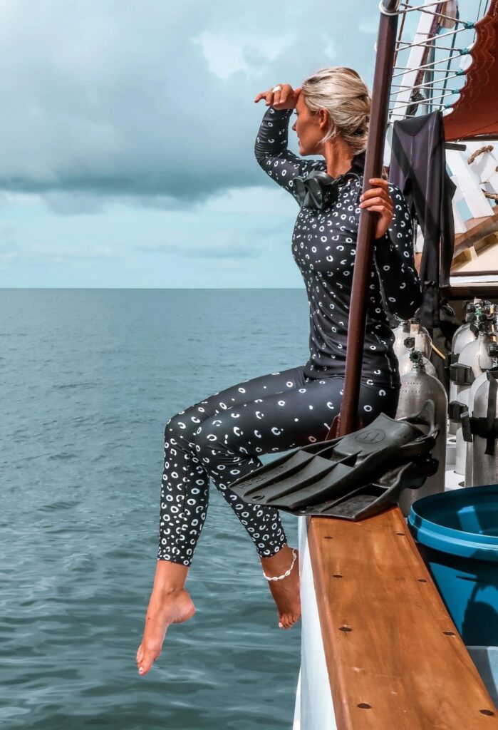 Charlotte sits on the side of a large scuba liveaboard looking out to the blue ocean. She is wearing a Blue Adaptation eagle ray set in black with a white print.