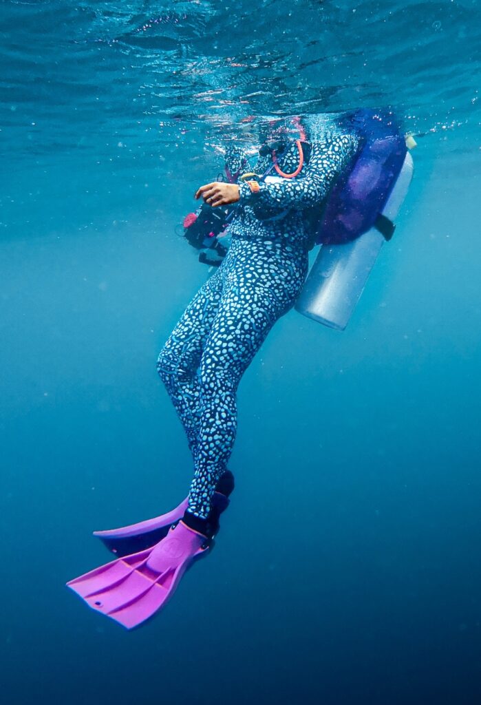 A woman scuba diver is just under the surface wearing a whale shark print set from the Girls that Scuba divewear collection. Her face isn't visible above the bright blue water. She has bright pink fins on and a purple BCD cover.