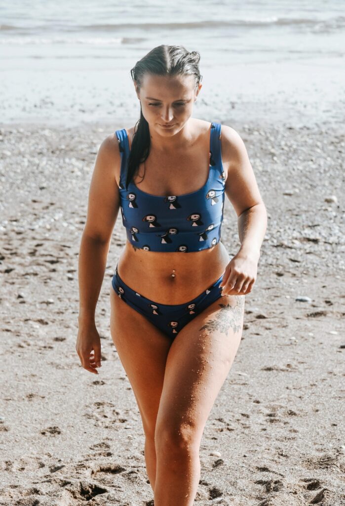 A woman walks towards the camera on a sandy beach. She is looking down and wearing a blue swimsuit with puffins on the print. 