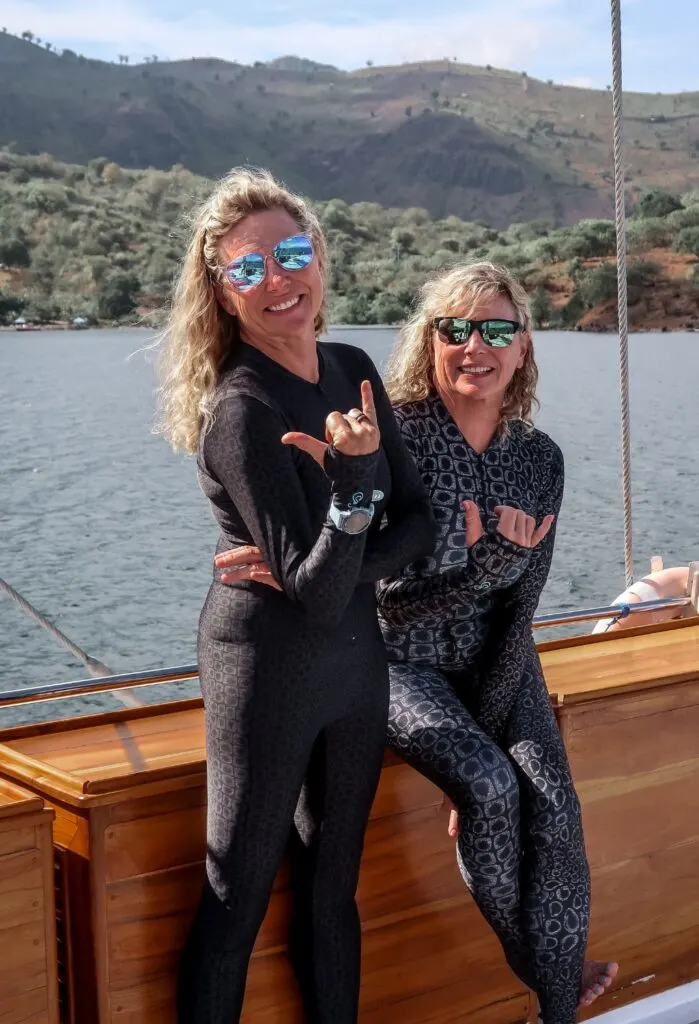 Two women smile at the camera and hold up their right hands in a "hang loose" or "shaka" hand signal with their thumbs and pinkie fingers extended. They are wearing matching black printed full dive suits from SlipIns. 