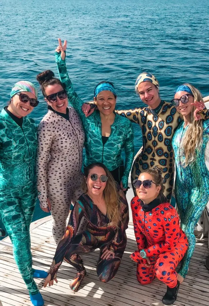 A group of 7 women smile at the camera wearing brightly coloured SlipIns suits, in blues, greens, yellows, and red. They are on the dive deck of a large boat with bright blue water to the back. 