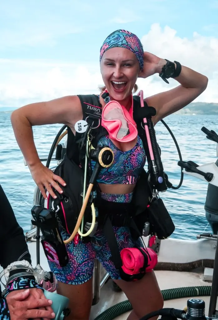 A woman scuba diver poses for the camera with one hand above her head and one on her hip. She is stood on a small boat, wearing a bright coral printed swimwear set, with a pink scuba mask to match. 
