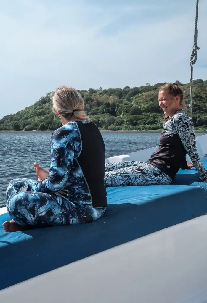 Two women sit on the side of a large boat laughing and smiling at one another. They are wearing matching black and printed women's divewear, one with whale sharks on the leggings and sleeves, and the other with an octopus-tentacle and flower print.