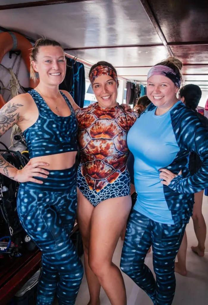 Three women smile at the camera wearing printed women's divewear, featuring whale shark prints and orange sea turtle inspired prints.