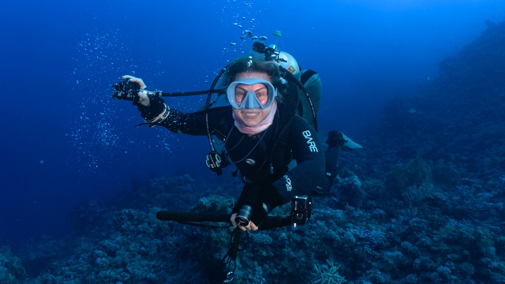 Scuba diver Krista is underwater, smiling at the camera whilst holding her regulator out of her mouth with her right hand. She is wearing a blue scuba mask and carrying a GoPro and dive light in her left hand. 