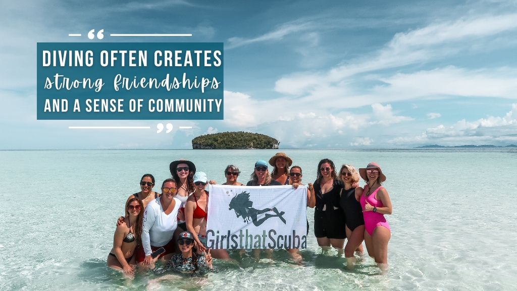 A group of women stand in shallow, crystal clear water with a small green island behind. They are smiling at the camera and holding a Girls that Scuba flag, with a symbol of a woman scuba diver silhouette.