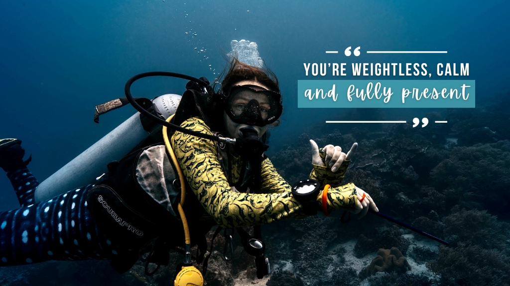 Amy is scuba diving and holding a "hang loose" hand symbol to the camera with her three middle fingers closed in and her thumb and pinky fingers extended. She is wearing a yellow rash vest with manta rays. Overlaid white text reads "You're weightless, calm, and fully present" 